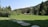 A lush green field is bordered by dense trees and bushes, leading to a cluster of rustic wooden cabins. The setting is backed by hills covered with thick pine forests under a clear sky. Stone pathways cross the vibrant grass, enhancing the idyllic, peaceful atmosphere.
