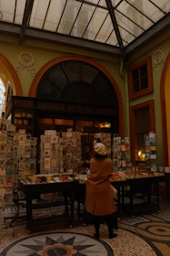 An indoor market or bookstore setup with numerous books and magazines displayed on tables and stands. A person wearing a beige coat and white beret stands in front of the display, browsing the items. The setting has a cozy, antique ambiance, with a high, glass-paneled ceiling and ornate architectural details.