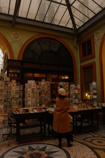 An indoor market or bookstore setup with numerous books and magazines displayed on tables and stands. A person wearing a beige coat and white beret stands in front of the display, browsing the items. The setting has a cozy, antique ambiance, with a high, glass-paneled ceiling and ornate architectural details.