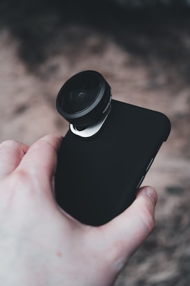 A hand holding a smartphone equipped with an external lens attachment, possibly for enhanced photography. The phone has a black case and the background is blurred.
