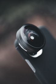 A close-up view of a camera lens affixed to a smartphone. The focus on the lens is sharp while the background is blurred, creating a depth of field effect. The brand 'SANDMARC' is visible on the lens mount. The overall composition highlights the sleek and modern design of the accessory.