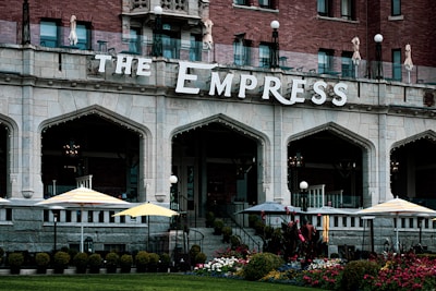 A grand brick and stone building with arched windows and decorative lighting. The facade features the name 'The Empress' prominently. There are several umbrellas and outdoor seating areas on a terrace, surrounded by neatly arranged flower beds and shrubs.