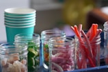 A row of glass jars filled with various colorful candies and snacks is arranged on a surface. To the left, there is a stack of teal paper cups. The jars contain an assortment of sweets such as green jelly candies, mixed color gummies, red licorice sticks, and different colored chewable treats.