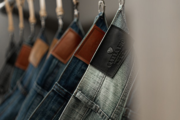 A row of denim jeans hanging from hangers, each featuring a distinct leather patch on the back waistband. The denim is in various shades of blue, with some patches showing brand labels.
