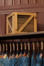A wooden crate labeled 'EXPORT' sits on a high shelf above a row of hanging blue jeans. The jeans are suspended by leather straps, and each pair features a leather tag near the waistband. The background consists of paneling in a brownish hue.