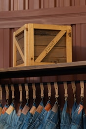 A wooden crate labeled 'EXPORT' sits on a high shelf above a row of hanging blue jeans. The jeans are suspended by leather straps, and each pair features a leather tag near the waistband. The background consists of paneling in a brownish hue.