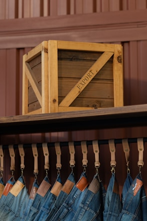 A wooden crate labeled 'EXPORT' sits on a high shelf above a row of hanging blue jeans. The jeans are suspended by leather straps, and each pair features a leather tag near the waistband. The background consists of paneling in a brownish hue.