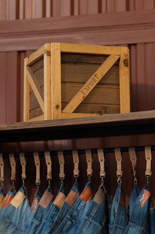 A wooden crate labeled 'EXPORT' sits on a high shelf above a row of hanging blue jeans. The jeans are suspended by leather straps, and each pair features a leather tag near the waistband. The background consists of paneling in a brownish hue.