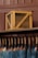 A wooden crate labeled 'EXPORT' sits on a high shelf above a row of hanging blue jeans. The jeans are suspended by leather straps, and each pair features a leather tag near the waistband. The background consists of paneling in a brownish hue.