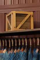 A wooden crate labeled 'EXPORT' sits on a high shelf above a row of hanging blue jeans. The jeans are suspended by leather straps, and each pair features a leather tag near the waistband. The background consists of paneling in a brownish hue.