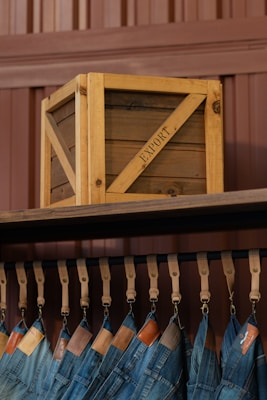 A wooden crate labeled 'EXPORT' sits on a high shelf above a row of hanging blue jeans. The jeans are suspended by leather straps, and each pair features a leather tag near the waistband. The background consists of paneling in a brownish hue.