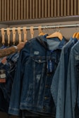 A hand is seen selecting a denim jacket from a rack of similar jackets. The rack is made of metal with wooden hangers supporting the jackets. The jackets have a worn, distressed look, and one of them has a tag attached.