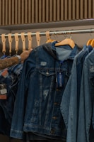 A hand is seen selecting a denim jacket from a rack of similar jackets. The rack is made of metal with wooden hangers supporting the jackets. The jackets have a worn, distressed look, and one of them has a tag attached.