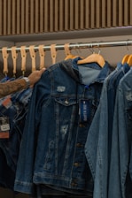 A hand is seen selecting a denim jacket from a rack of similar jackets. The rack is made of metal with wooden hangers supporting the jackets. The jackets have a worn, distressed look, and one of them has a tag attached.