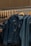 A hand is seen selecting a denim jacket from a rack of similar jackets. The rack is made of metal with wooden hangers supporting the jackets. The jackets have a worn, distressed look, and one of them has a tag attached.