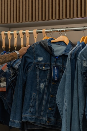 A hand is seen selecting a denim jacket from a rack of similar jackets. The rack is made of metal with wooden hangers supporting the jackets. The jackets have a worn, distressed look, and one of them has a tag attached.