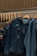 A hand is seen selecting a denim jacket from a rack of similar jackets. The rack is made of metal with wooden hangers supporting the jackets. The jackets have a worn, distressed look, and one of them has a tag attached.
