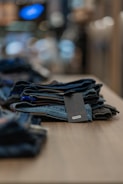 A neatly folded stack of denim jeans is placed on a wooden table. The focus is on the jeans, with a blurred background that suggests an indoor setting, possibly a store. A label with the brand name is visible on the top pair of jeans.