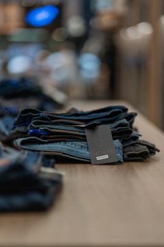 A neatly folded stack of denim jeans is placed on a wooden table. The focus is on the jeans, with a blurred background that suggests an indoor setting, possibly a store. A label with the brand name is visible on the top pair of jeans.