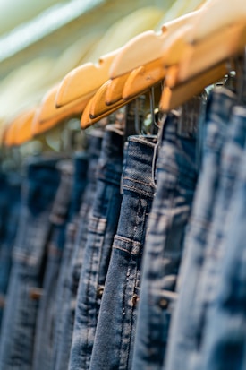 A row of blue denim jeans hanging on wooden hangers, closely aligned and creating a repetitive pattern. The focus is on the textured fabric and the metal details of the jeans.