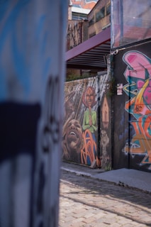 Colorful graffiti art adorns the walls of an urban alley. The artwork features vibrant and expressive faces, with abstract shapes and vivid colors, adding an artistic and lively touch to the rustic surroundings.