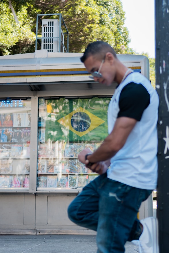 A person wearing sunglasses and a white and black t-shirt leans against a pole on a street. Behind them, a newsstand displays a Brazilian flag along with various magazines and publications. Lush green trees surround the scene, hinting at a sunny day.