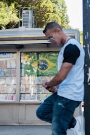 A person wearing sunglasses and a white and black t-shirt leans against a pole on a street. Behind them, a newsstand displays a Brazilian flag along with various magazines and publications. Lush green trees surround the scene, hinting at a sunny day.