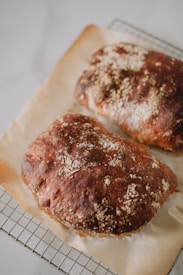 Two loaves of rustic bread with a dark, crusty surface are resting on a piece of parchment paper placed over a wire cooling rack.