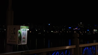 A green public SOS phone is situated on a bridge at night, illuminated against a dark backdrop. City lights are faintly visible in the distance, and the railing of the bridge has blue lighting accents.