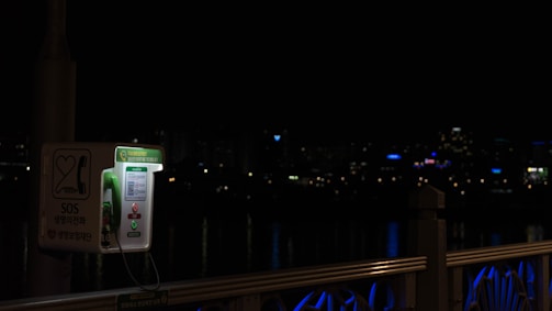 A green public SOS phone is situated on a bridge at night, illuminated against a dark backdrop. City lights are faintly visible in the distance, and the railing of the bridge has blue lighting accents.