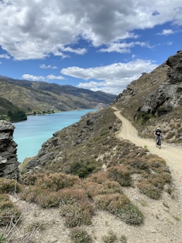 A cyclist rides along a rugged dirt path that winds through hilly terrain by a picturesque turquoise lake, surrounded by rocky cliffs and sparse vegetation. Fluffy white clouds dot the bright blue sky, creating a serene yet adventurous atmosphere.
