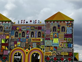 A colorful, eclectic facade featuring a mix of geometric shapes and varied patterns with numerous words and phrases like 'LOVE', 'GRIT', 'FREEDOM', and 'HOPE'. The design incorporates a mix of bright and bold colors, giving it a striking visual appeal. The intricate layout is reminiscent of a playful, yet meaningful, mural or installation art piece.
