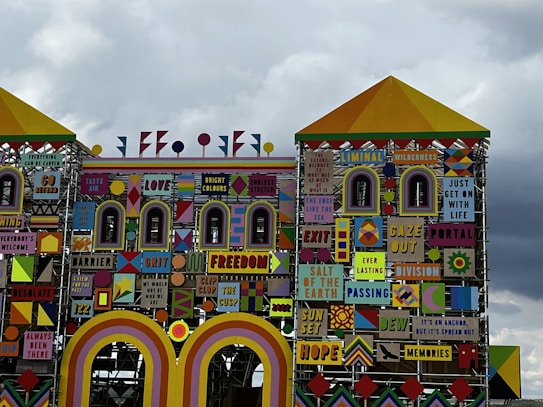 A colorful, eclectic facade featuring a mix of geometric shapes and varied patterns with numerous words and phrases like 'LOVE', 'GRIT', 'FREEDOM', and 'HOPE'. The design incorporates a mix of bright and bold colors, giving it a striking visual appeal. The intricate layout is reminiscent of a playful, yet meaningful, mural or installation art piece.