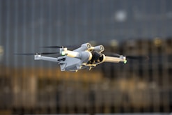 A white drone with black propellers is captured mid-flight against a blurred background of tall glass buildings. The drone has green and yellow lights on its arms and a camera attached to its main body.