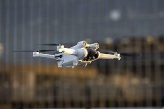 A white drone with black propellers is captured mid-flight against a blurred background of tall glass buildings. The drone has green and yellow lights on its arms and a camera attached to its main body.