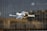 A white drone with black propellers is captured mid-flight against a blurred background of tall glass buildings. The drone has green and yellow lights on its arms and a camera attached to its main body.