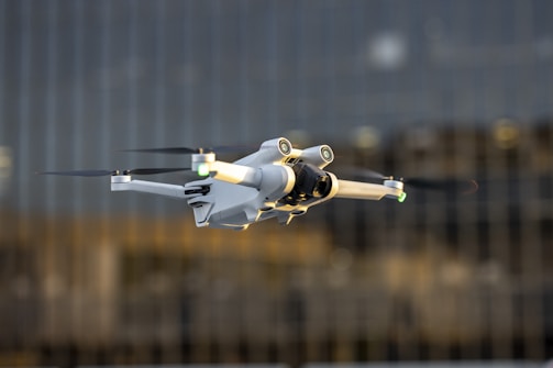 A white drone with black propellers is captured mid-flight against a blurred background of tall glass buildings. The drone has green and yellow lights on its arms and a camera attached to its main body.