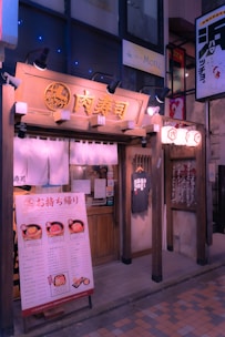 A quaint Japanese restaurant facade with traditional wooden architecture, hanging fabric banners, and illuminated signage. There is a menu board displaying food options and prices at the entrance, along with a T-shirt on display. Lanterns hang near the doorway, adding to the inviting and cozy atmosphere.