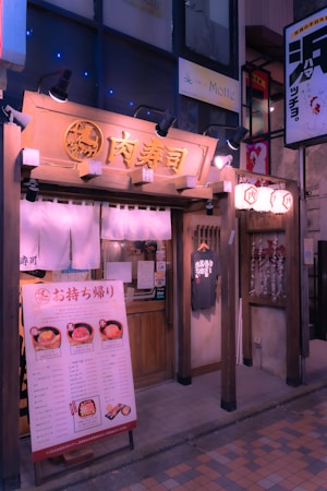A quaint Japanese restaurant facade with traditional wooden architecture, hanging fabric banners, and illuminated signage. There is a menu board displaying food options and prices at the entrance, along with a T-shirt on display. Lanterns hang near the doorway, adding to the inviting and cozy atmosphere.