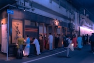 A busy street scene in an urban area features a group of people queuing outside a restaurant. The exterior is styled with wooden panels, and the soft glow of neon lights casts a blue and purple hue over the scene. People are dressed in varying styles, suggesting a casual evening atmosphere.