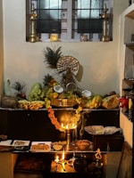 A traditional altar setup with two tall brass lamps flanking a variety of fruits, flowers, and offerings displayed on a dark platform. There is an assortment of fruits including bananas, mangoes, and coconuts, alongside sweets and nuts arranged neatly. Peacocks feathers and a central deity statue provide a cultural and spiritual element. Soft lighting from oil lamps adds a warm glow to the arrangement, creating an atmosphere of reverence.