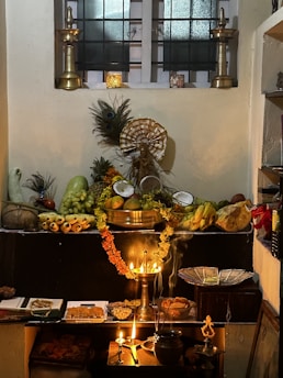 A traditional altar setup with two tall brass lamps flanking a variety of fruits, flowers, and offerings displayed on a dark platform. There is an assortment of fruits including bananas, mangoes, and coconuts, alongside sweets and nuts arranged neatly. Peacocks feathers and a central deity statue provide a cultural and spiritual element. Soft lighting from oil lamps adds a warm glow to the arrangement, creating an atmosphere of reverence.