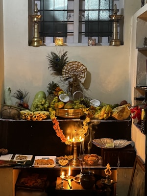 A traditional altar setup with two tall brass lamps flanking a variety of fruits, flowers, and offerings displayed on a dark platform. There is an assortment of fruits including bananas, mangoes, and coconuts, alongside sweets and nuts arranged neatly. Peacocks feathers and a central deity statue provide a cultural and spiritual element. Soft lighting from oil lamps adds a warm glow to the arrangement, creating an atmosphere of reverence.