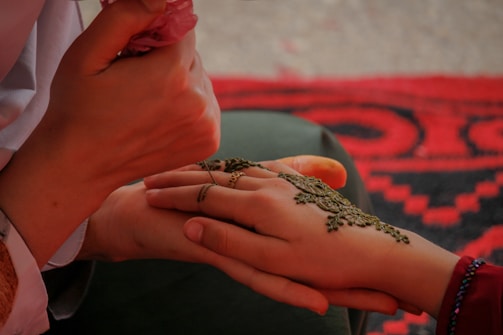 A person is applying intricate henna designs on another person's hand. The focus is on the detailed work being done, with a mixture of steady and delicate lines forming floral patterns. The setting includes a vibrant, patterned fabric in the background, adding a cultural ambiance to the scene.