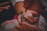 A henna artist applies intricate floral and geometric designs on a person's palm using a henna applicator. The hand rests on a colorful, patterned cloth, and there is a serene and detailed focus on the artistry and cultural tradition of henna application.