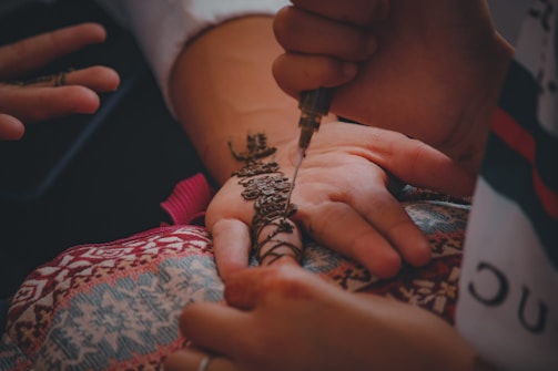 A henna artist applies intricate floral and geometric designs on a person's palm using a henna applicator. The hand rests on a colorful, patterned cloth, and there is a serene and detailed focus on the artistry and cultural tradition of henna application.