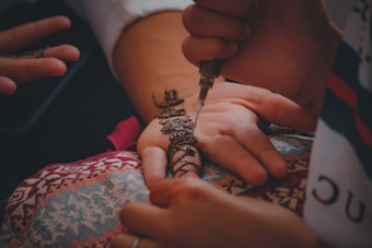 A henna artist applies intricate floral and geometric designs on a person's palm using a henna applicator. The hand rests on a colorful, patterned cloth, and there is a serene and detailed focus on the artistry and cultural tradition of henna application.