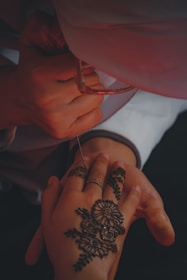 A person's hand is being adorned with intricate henna designs. The artwork is detailed with floral patterns and swirls. The hand is gently held by another person who is applying the henna using a cone. Soft lighting casts a warm glow over the scene, accentuating the artistry and careful application.