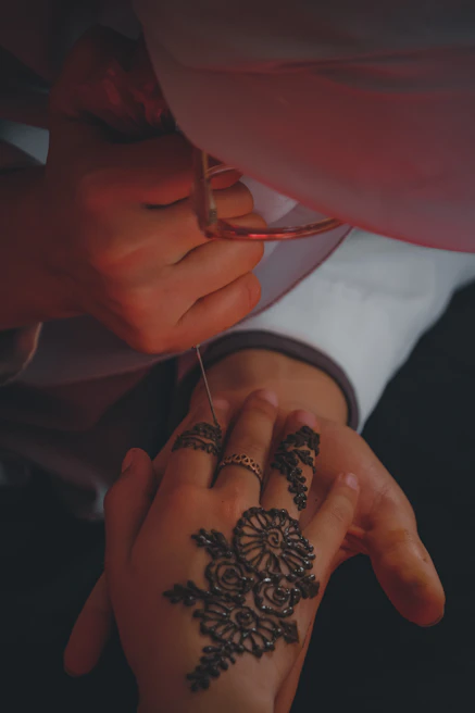 Close-up of a skilled hand applying intricate henna designs on a client's hand in soft lighting.