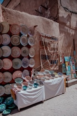 A vibrant street market display featuring an array of intricately designed ceramic plates and bowls arranged on two tables. To the left, numerous plates are mounted on a wall, showcasing various colorful patterns. A woven textile hangs next to them, and to the right, small framed artworks are visible on the ground.